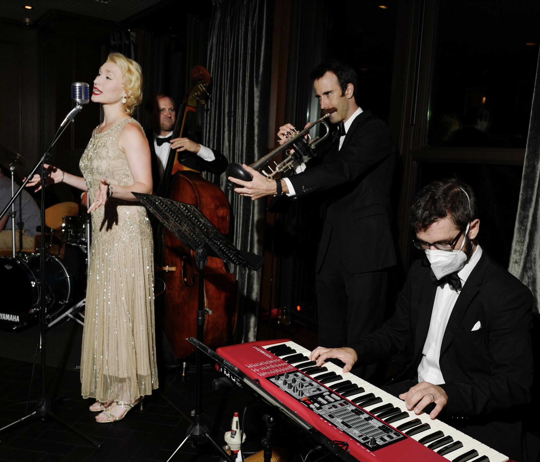 An image of a jazz quarter performing, including a woman singing, man playing piano, another man playing trumpet, and a third man playing a string instrument.