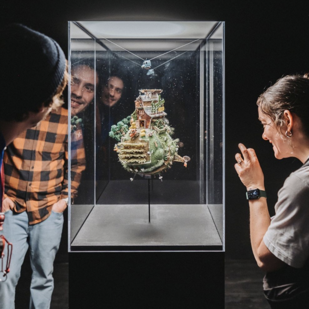 An image of three people looking at a miniature piece of art from the Small Is Beautiful Exhibit. The artwork they are looking at is a small globe with a house, farm, and other natural elements.